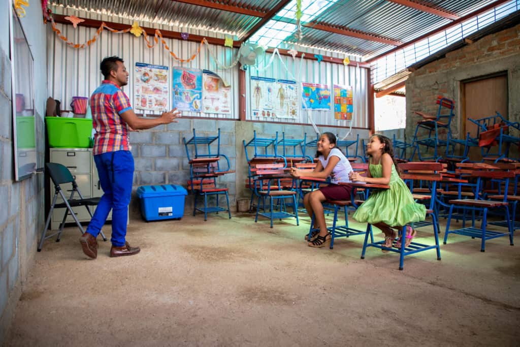 Girl wearing a green dress and pink sandals and is sitting with a friend, wearing a white shirt, a red skirt and black sandals. They are sitting in one of the newly constructed classrooms of a Compassion center. The project tutor is in the front of the classroom, wearing a red plaid shirt and blue pants. 