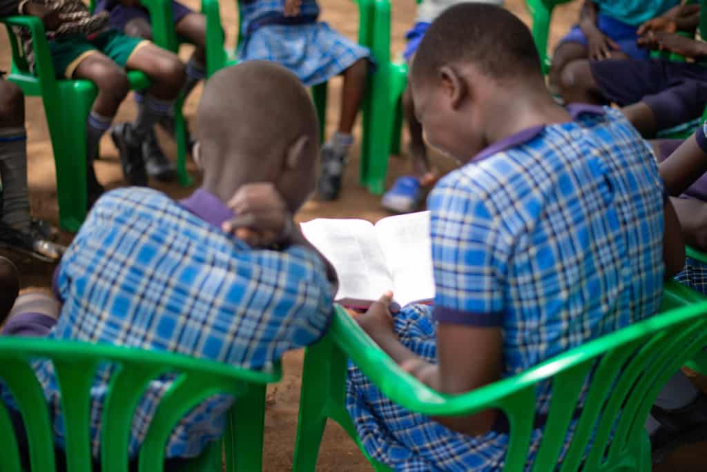 Rita and several other children are sitting outside in green plastic chairs. They are reading the Bible together and they are all wearing their school uniforms, blue plaid dresses.