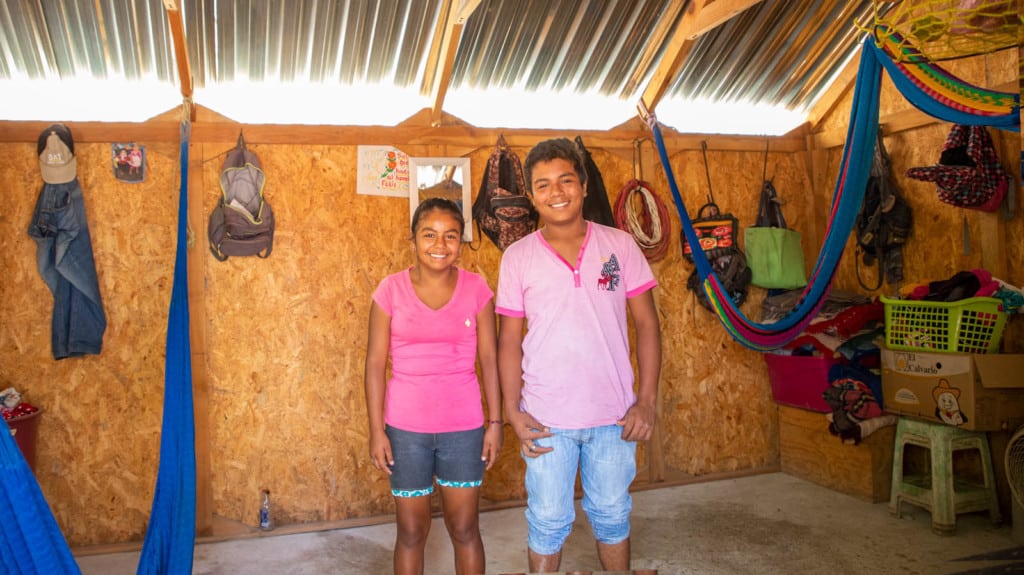 Girl wearing a dark pink shirt and her brother is wearing a light pink shirt. They are standing in their home between the hammocks they use during the day and at night to sleep.