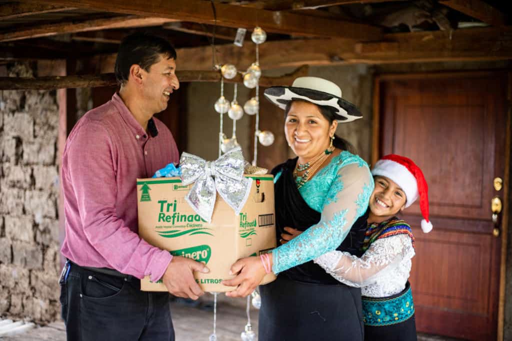 Miguel is giving a food box to Jenny while Joselin hugs her mother. They are wearing traditional clothes. There are Christmas lights behind them.