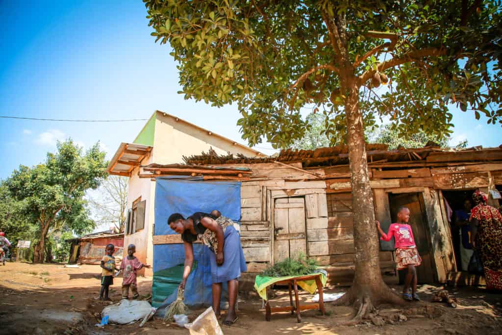 A woman in a black shirt and blue skirt, is sweeping the ground outside her wood house. She is carrying an infant in a sling on her back.