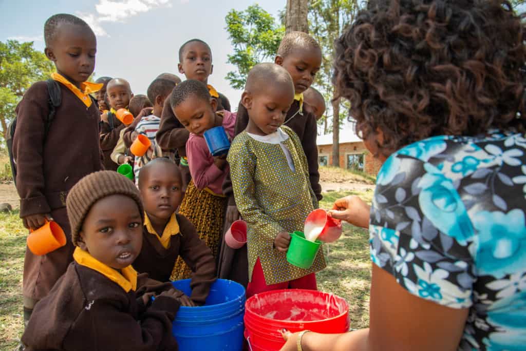 Kindergarten students at Ekenywa Pre and Primary English Medium School are in a line being served porridge by their teacher. The students are wearing brown sweaters and are outside.