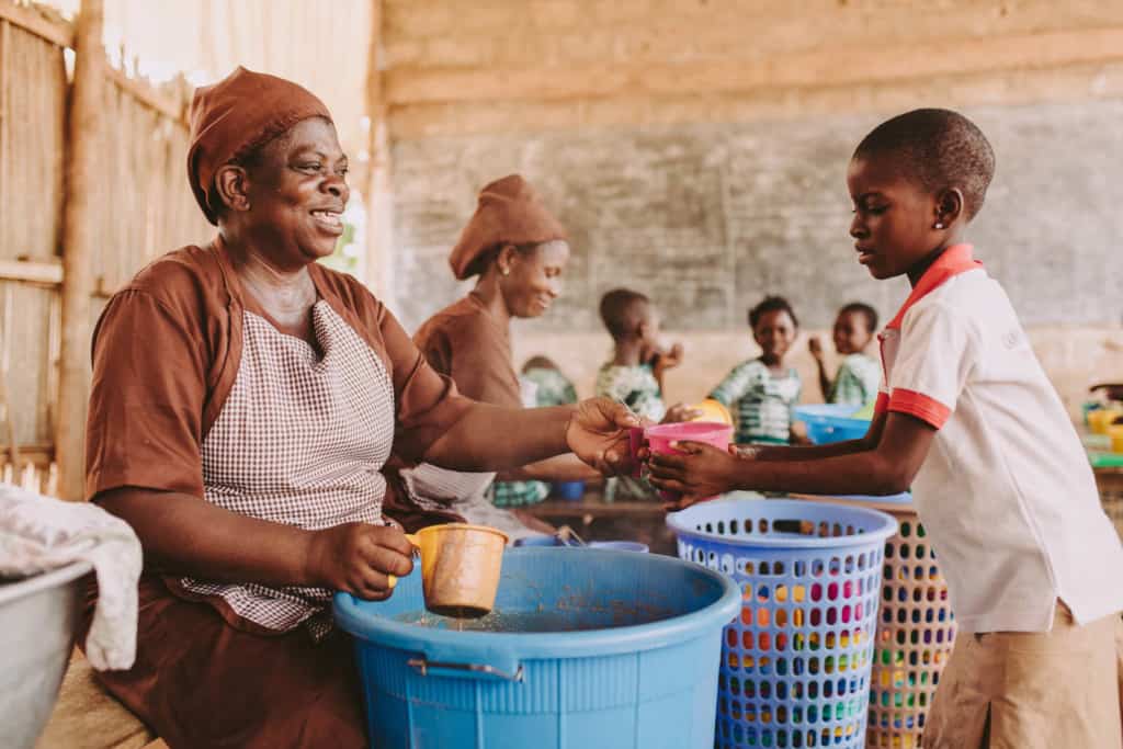 A girl child wearing school uniform center white shirt with a brown skirt holds her hands out standing in line to receive cups of nutritious food meals lunch from colorful plastic bowls cups handed to them from a Compassion staff adult woman wearing brown dress apron and scarf on her head. 