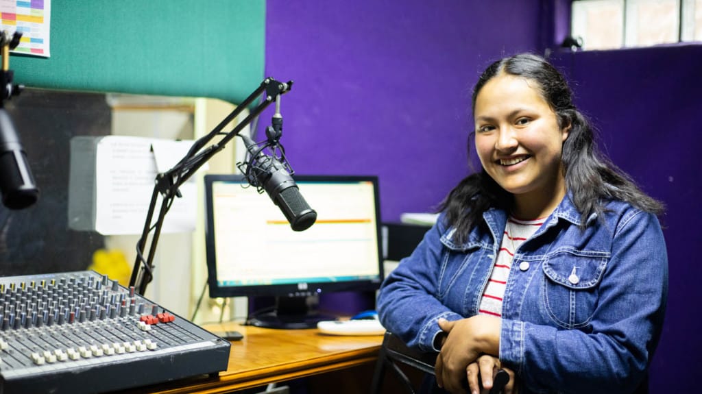 Itzel is wearing a jean jacket and is sitting in the radio studio. There is a microphone and a sound board beside her.