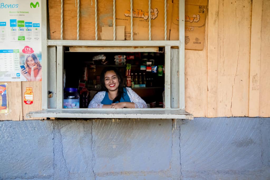 Woman standing inside her house, and leaning against the edge of the window from which they sell food, canned goods, candies and drinks like sodas and orange juice. She is wearing a blue shirt and a white cardigan while looking directly at the camera and smiling. 