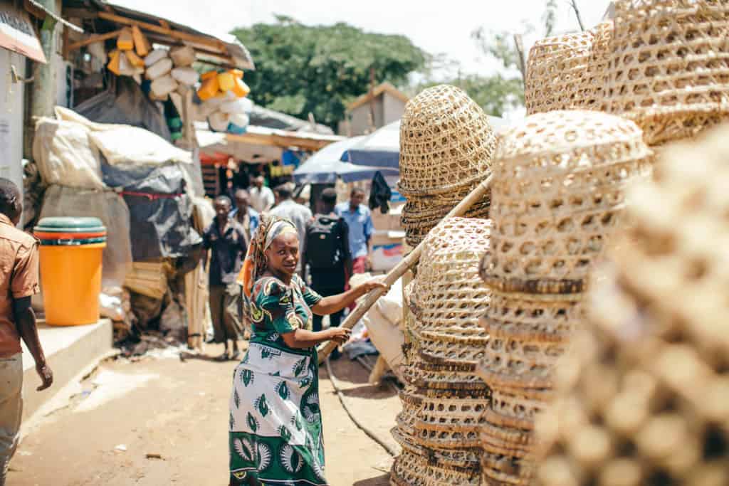 A woman in a green and white dress with a scarf on her head holds a stick over stacks of woven baskets at a market. There are more people walking in the background.