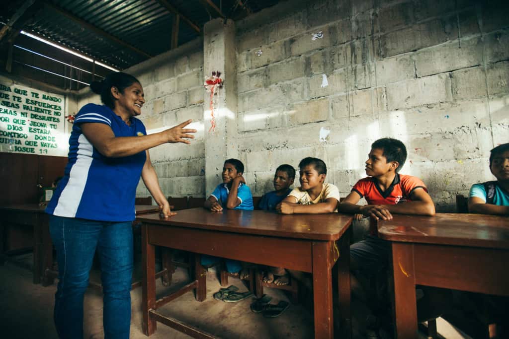 A woman in a blue and white shirt and blue jeans stands in front of a group of boys sitting at brown wood desks. There is a gray cinder block, cement wall behind them.