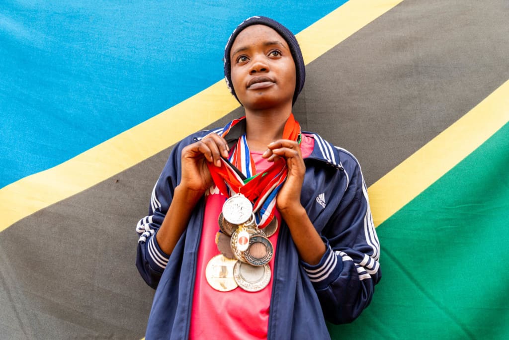 Clenzeensia, in a hot pink shirt and navy blue jacket and cap, is standing in front of the Tanzanian flag with her medals hanging from ribbons around her neck.