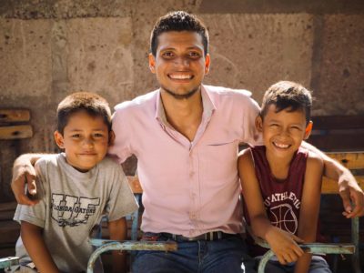 A pastor of a church in Nicaragua wears a pink button-down shirt and smiles as he sits next to two smiling young boys