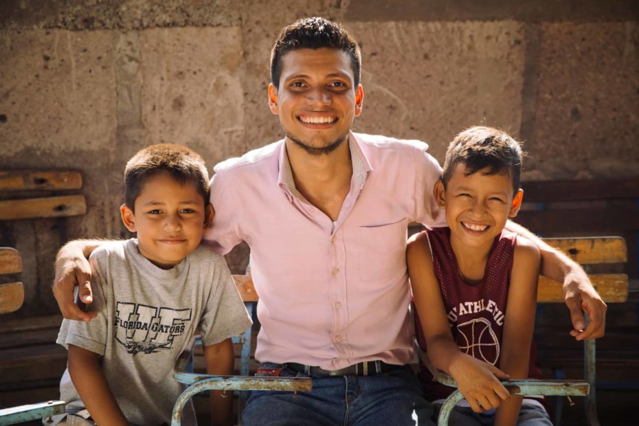 A pastor of a church in Nicaragua wears a pink button-down shirt and smiles as he sits next to two smiling young boys