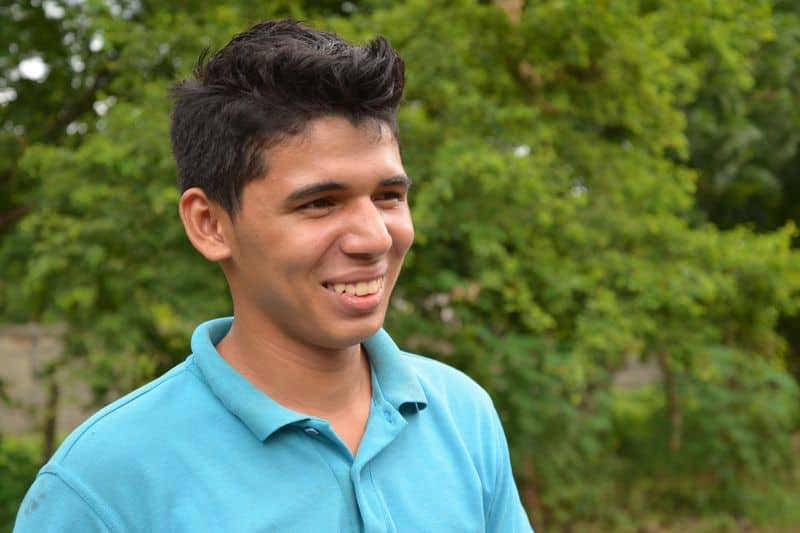 Junior, a teenage boy, smiles. He is wearing a blue polo shirt. Green trees are behind him