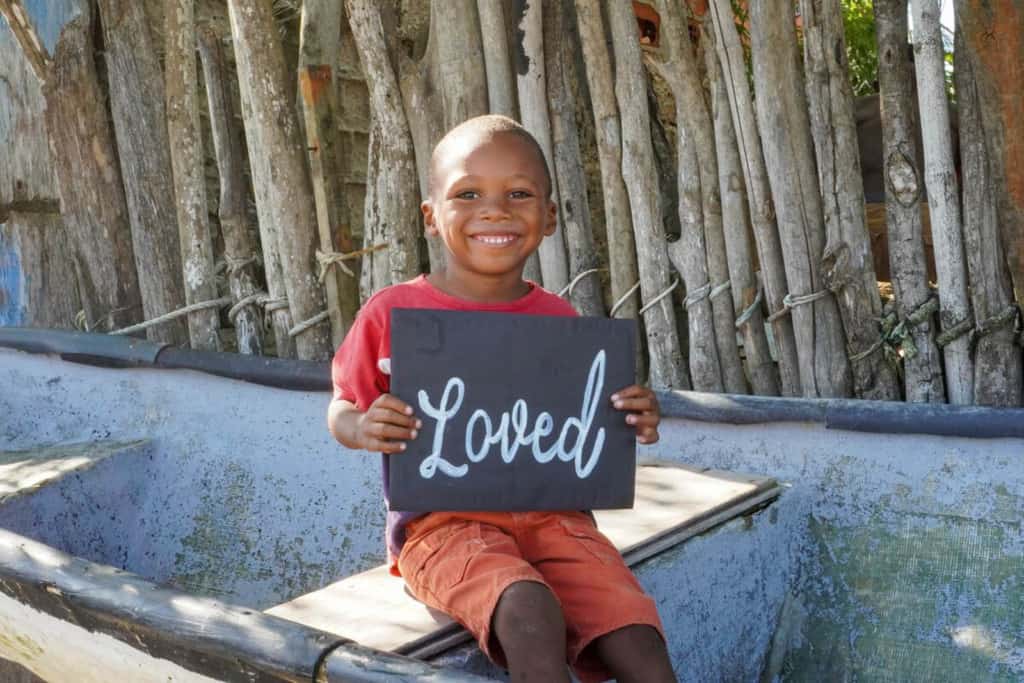 A boy is in his community and is sitting on a small ship on the beach. He smiles while holding a poster that says he is “loved” at the Compassion center.