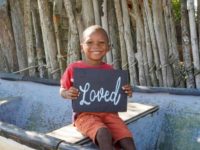 A boy is in his community and is sitting on a small ship on the beach. He smiles while holding a poster that says he is “loved” at the Compassion center.