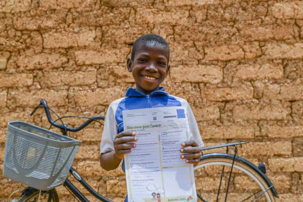 Boy wearing a blue shirt with white sleeves. He is standing in front of his bike and is holding a letter he received from his sponsor. Behind him is a tan brick wall.