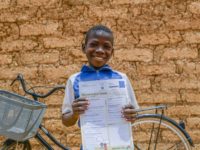 Boy wearing a blue shirt with white sleeves. He is standing in front of his bike and is holding a letter he received from his sponsor. Behind him is a tan brick wall.