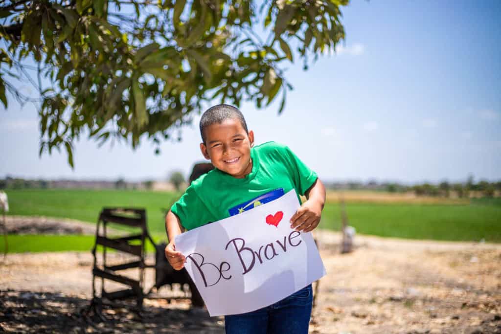 Gino is wearing a green shirt and jeans. He is standing outside with a rice field behind him and he is holding a sign that says Be Brave.