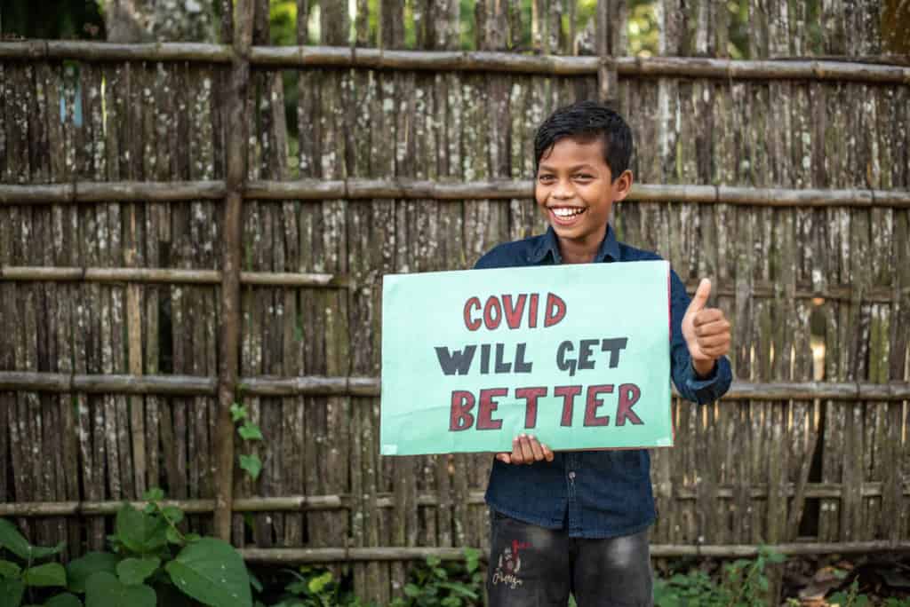 Bikash is wearing a navy blue shirt and jeans. He is holding a green sign that says, "COVID will get better." He is standing in front of a wooden fence and is giving a thumbs up sign.