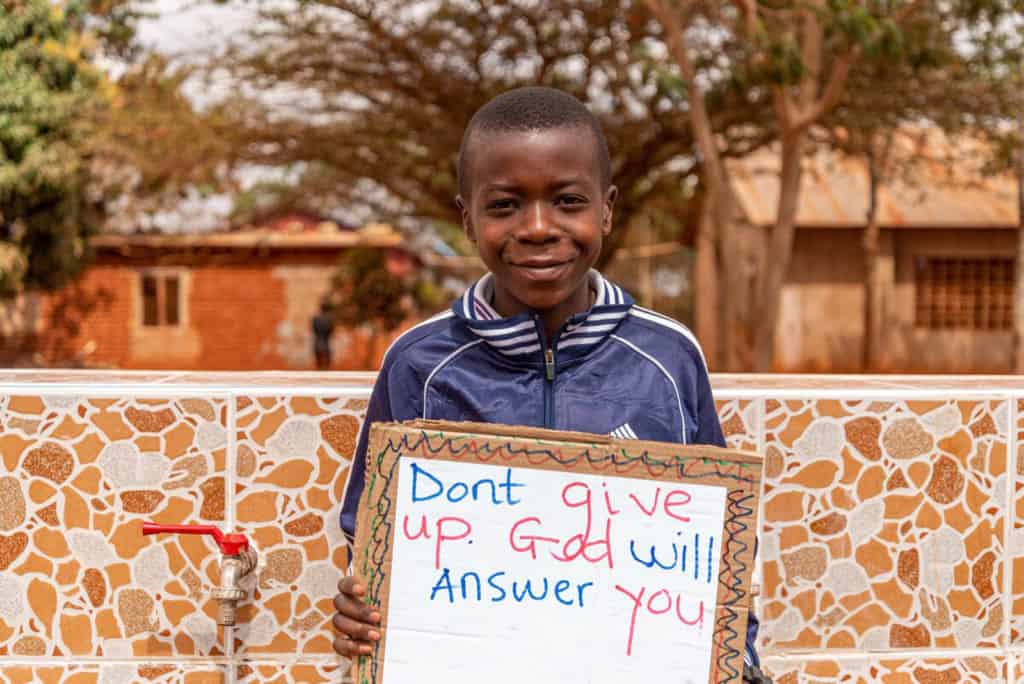 John is wearing a blue jacket and is holding up a sign that says, "Don't give up. God will answer you."