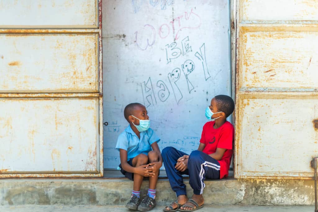 Charles is wearing a blue shirt. He is sitting with his friend, Emmanuel, wearing a red shirt and blue pants. They are sitting in front of a sign that says, "Don't worry, be happy." Both boys are wearing face masks.