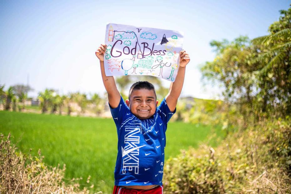 Antonio is wearing a blue shirt and red shorts. He is standing outside with a rice field behind him and he is holding up a sign that says God Bless You.