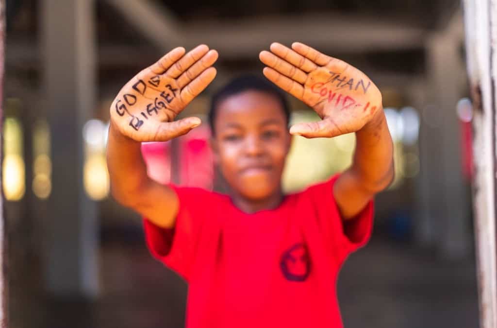 Emmanuel is wearing a red shirt. On his hands is written "God is bigger than COVID-19." He is holding his hands out toward the camera.