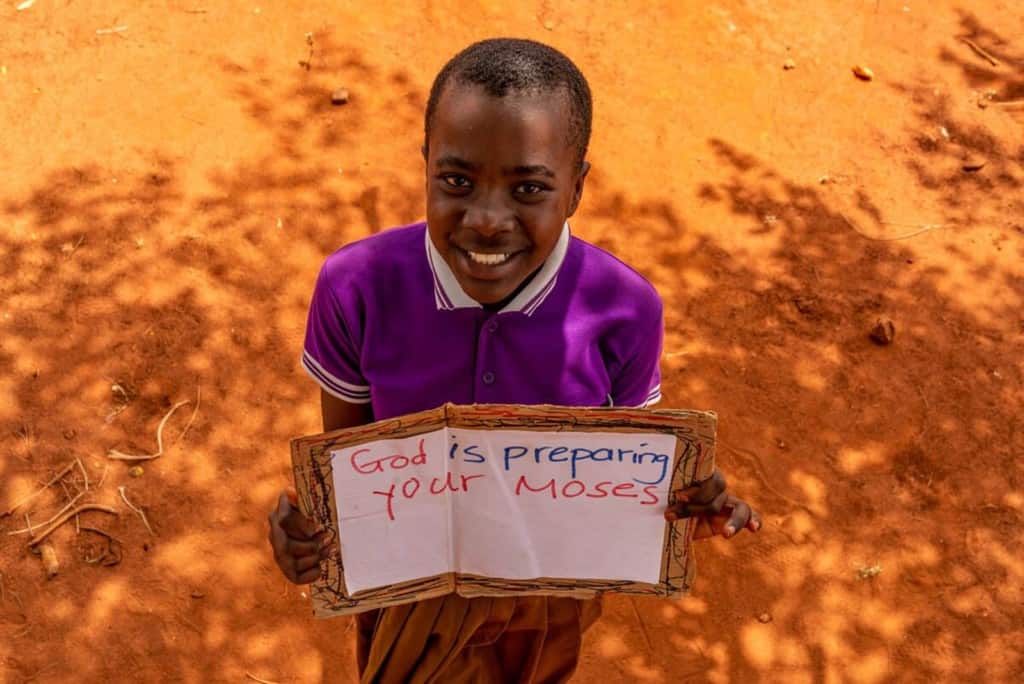 Zawadi is wearing a purple shirt and a tan skirt. She is standing outside and is holding a sign that says, "God is preparing your Moses."