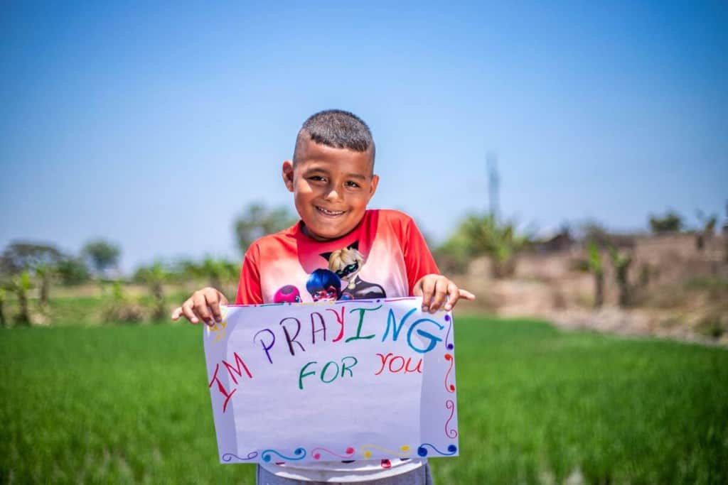 Caleb is weairng a white shirt with a pattern on the front and red sleeves. He is standing outside with a rice field behind him and is holding up a sign that says I'm Praying For You.
