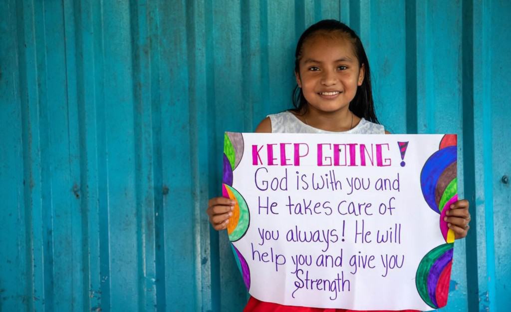 Fatima is holding up a sign that says: Keep going, God is with you and He takes care of you always! He will help you and give you strength. She is standing in front of a blue metal wall at the Compassion center.