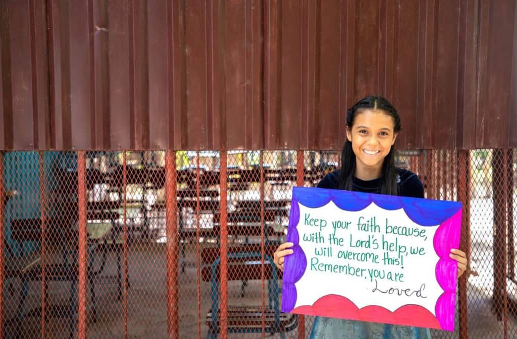 Josebeth is wearing a black shirt and is holding up a sign that says: keep your faith because, with the Lord’s help, we will overcome this! Remember, you are loved! She is standing in front of a brown wall at the Compassion center.
