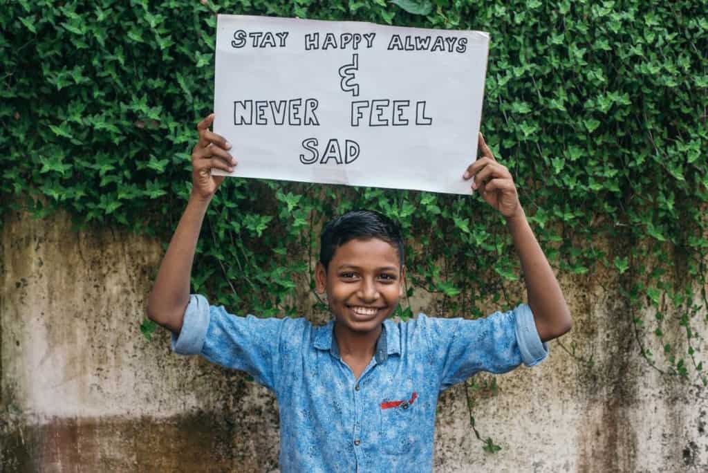Tawhid is wearing a blue shirt and jeans. He is holding up a sign that says, "Stay happy always and never feel sad." Behind him is a concrete wall covered partially with ivy.
