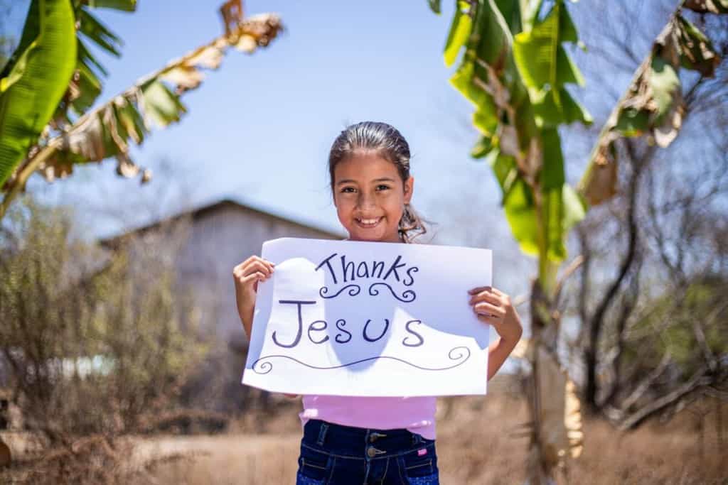 Haydee is wearing a pink shirt and jeans. She is standing outside her home and is surrounded by banana trees and is holding a sign that says Thanks Jesus.