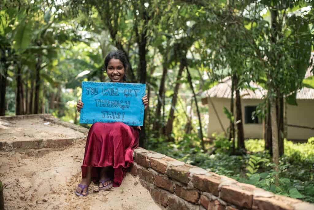 Sumona is wearing a dress with a floral bodice and a red skirt. She is sitting down on a short brick wall and is holding a blue sign that says, "You will get through this." There are trees and a house behind her.
