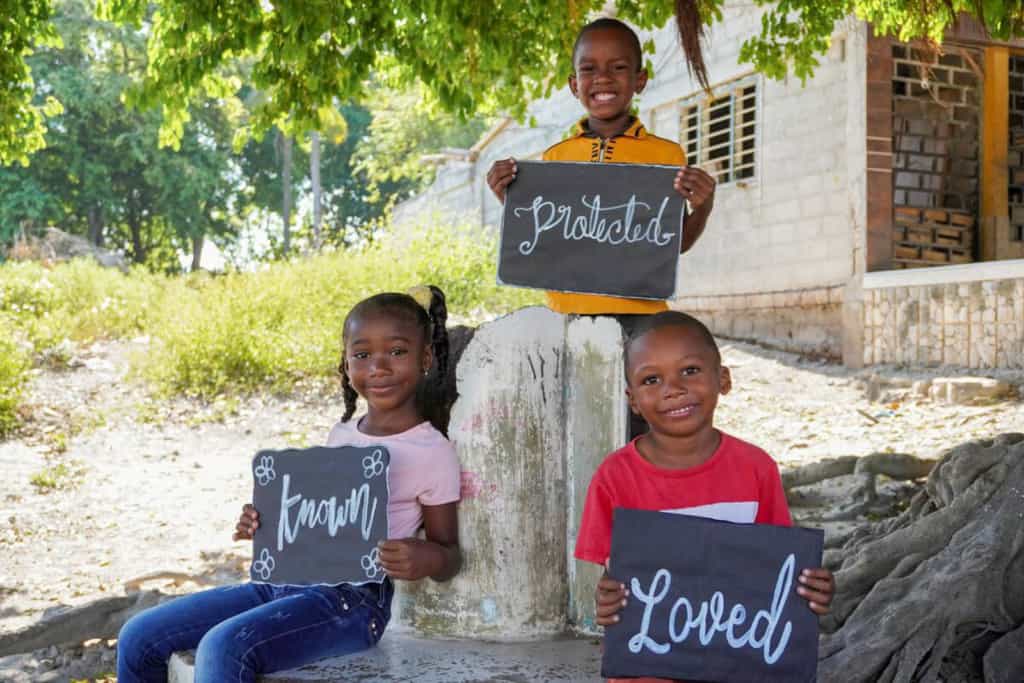 Three children in their community on the island where they live. They are holding posters to show they are known, loved, and protected at their Compassion center.