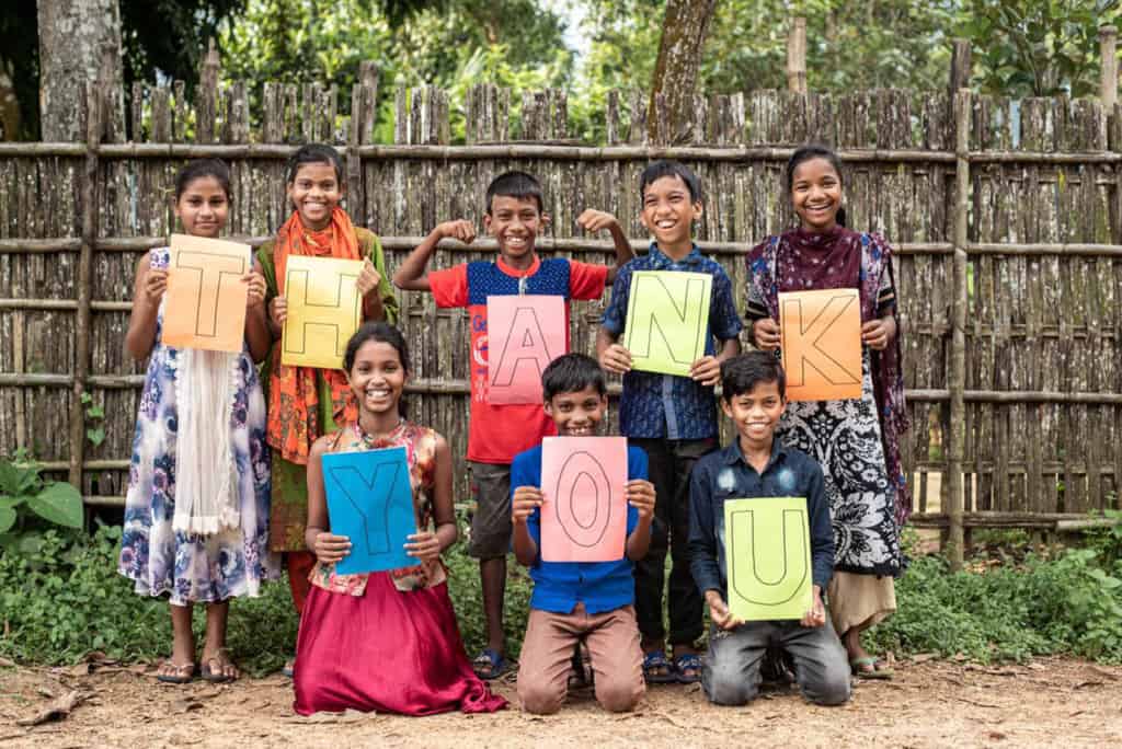 A group of children are holding up letters on colorful paper that spell out Thank you. They are in front of a wooden fence.