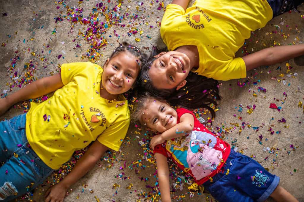 Ashley and Emily are wearing yellow shirts and jeans. They are laying down with their younger sister, Dara, wearing a red shirt and jean shorts. THey are laying in confetti with their heads together.