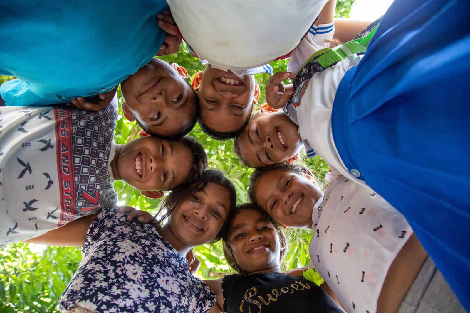 A group of beneficiary children are standing outside with their heads together and are looking down at the camera.