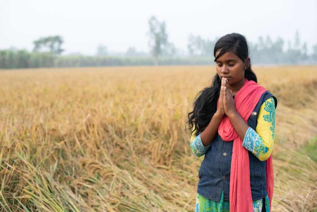 Girl in a yellow, green and blue dress, blue vest, and red, pink scarf, stands in a field of tall grass with her eyes closed and hands held together in front of her, praying. There are trees in the background.