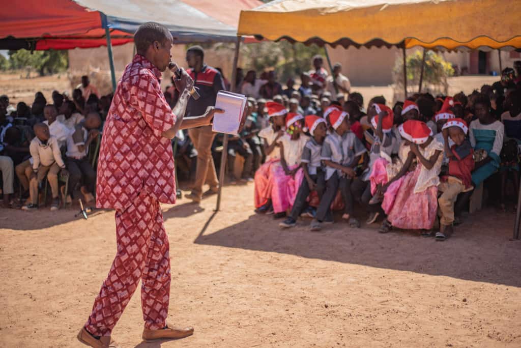 The pastor, in red, is preaching the gospel to the beneficiaries and guests at the ceremony outside the church compound. He is holding a Bible, a notebook and a microphone. A group of children wearing red and white Santa hats are sitting in the shade under a tent.