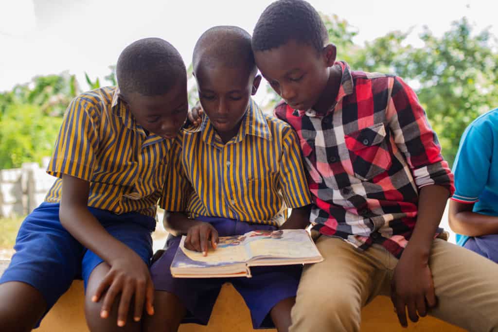 Children in purple, blue and yellow uniforms and a black and red shirt are sitting together reading an illustrated Bible.