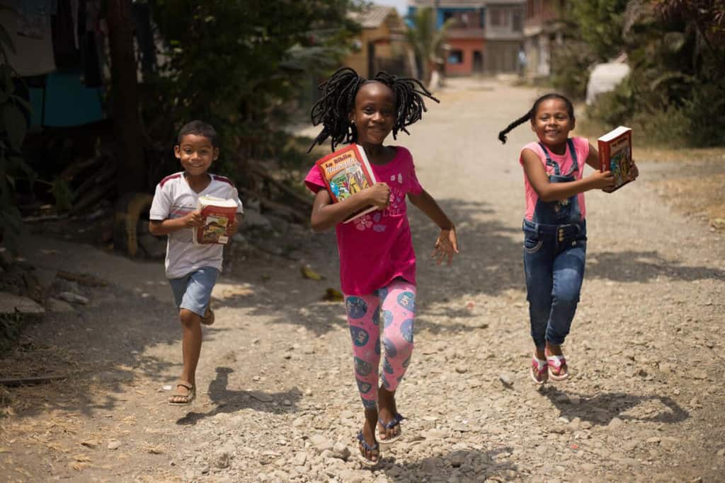 Samuel, in a white shirt, María Salome, in a fuchsia t-shirt, and Maria Angelica, in denim overalls, are running home along a dirt, gravel road while holding the Illustrated Bibles they received at the project.
