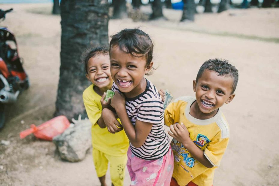 Girl, in a pink and black shirt, is standing on the sand laughing with another girl and a boy, both wearing yellow. Julenda is not part of the Child Sponsorship Program. There are trees in the background.