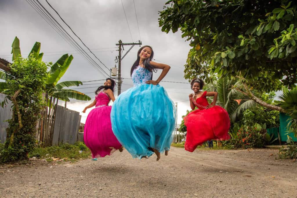 Lesly, wearing a blue dress, is with her friends, Ledis, wearing a pink dress, and Celena, wearing a red dress. They are all jumping in the air outside the project. There are trees behind them.