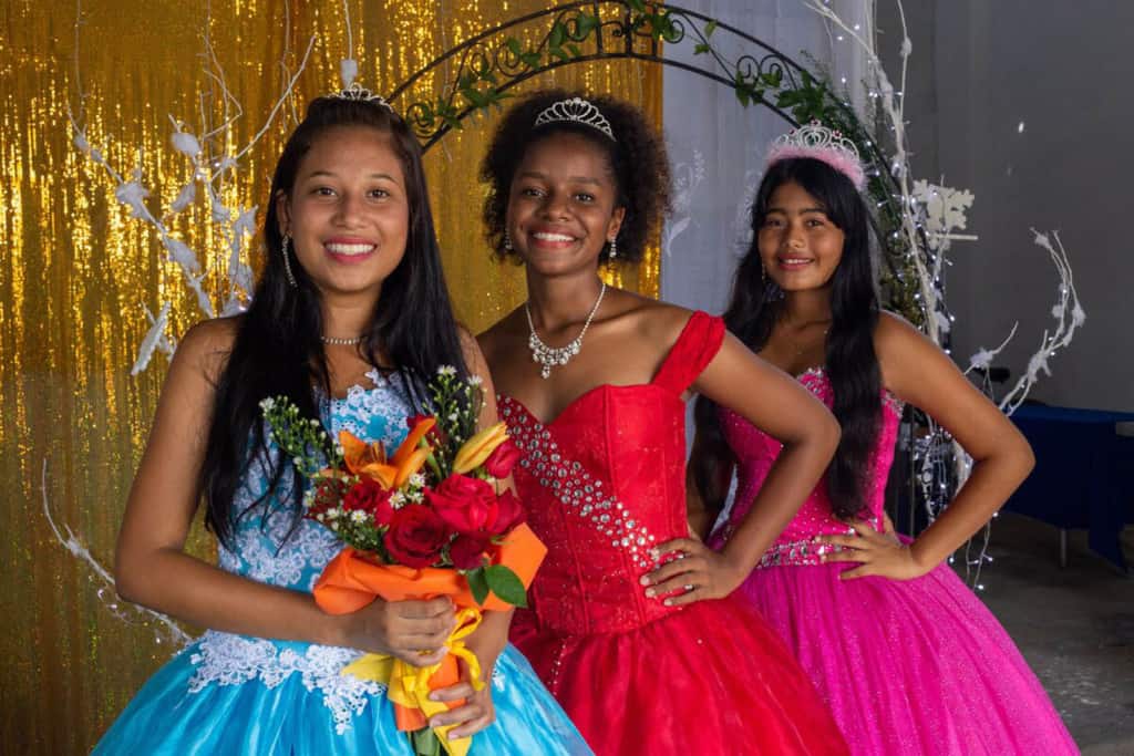 Lesly, wearing a blue dress, Celena, wearing a red dress, and Ledis, wearing a pink dress are standing together at the Compassion project. There is a gold curtain behind them.