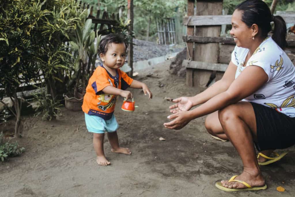 Toddler is wearing light blue shorts and an orange shirt. His mother is wearing black shorts and a white shirt. She is kneeling down with her arms outstretched helping him walk. Behind them is a fence.
