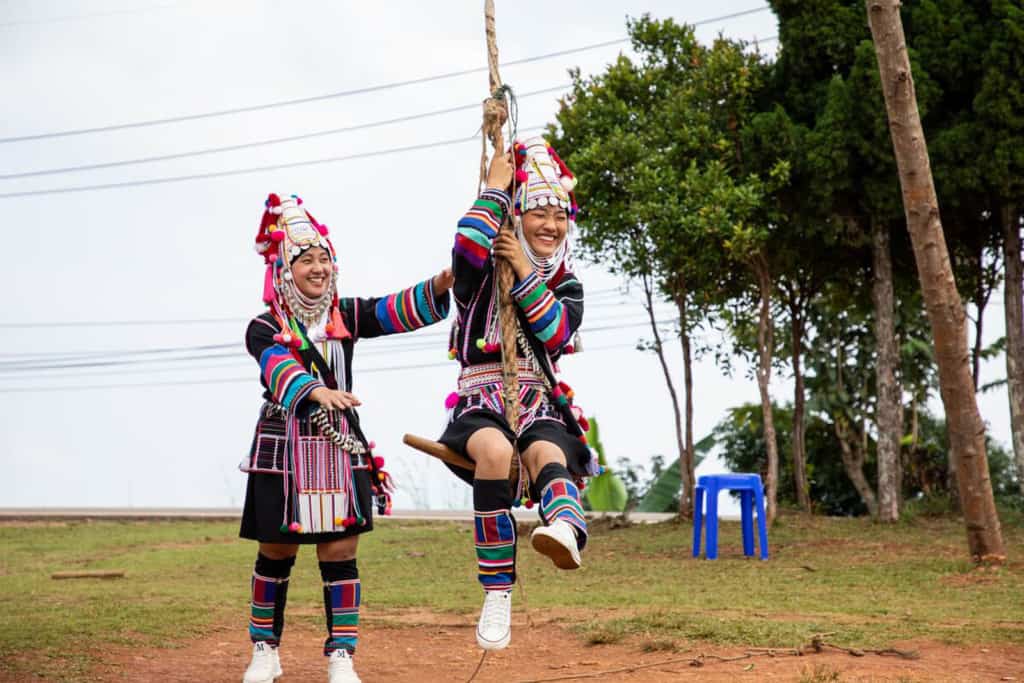 Abo (left, standing) is pushing Kanyaporn (right) on an Akha traditional celebration swing. They are wearing their traditional clothing and there are trees in the background.