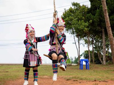 Abo (left, standing) is pushing Kanyaporn (right) on an Akha traditional celebration swing. They are wearing their traditional clothing and there are trees in the background.