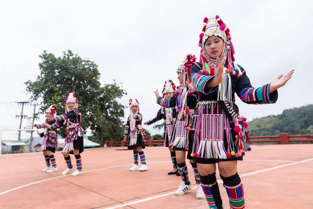 Abo (right) practicing the Akha traditional female dance with her friends for an upcoming celebration event. They are outside wearing their traditional clothing with trees in the background.