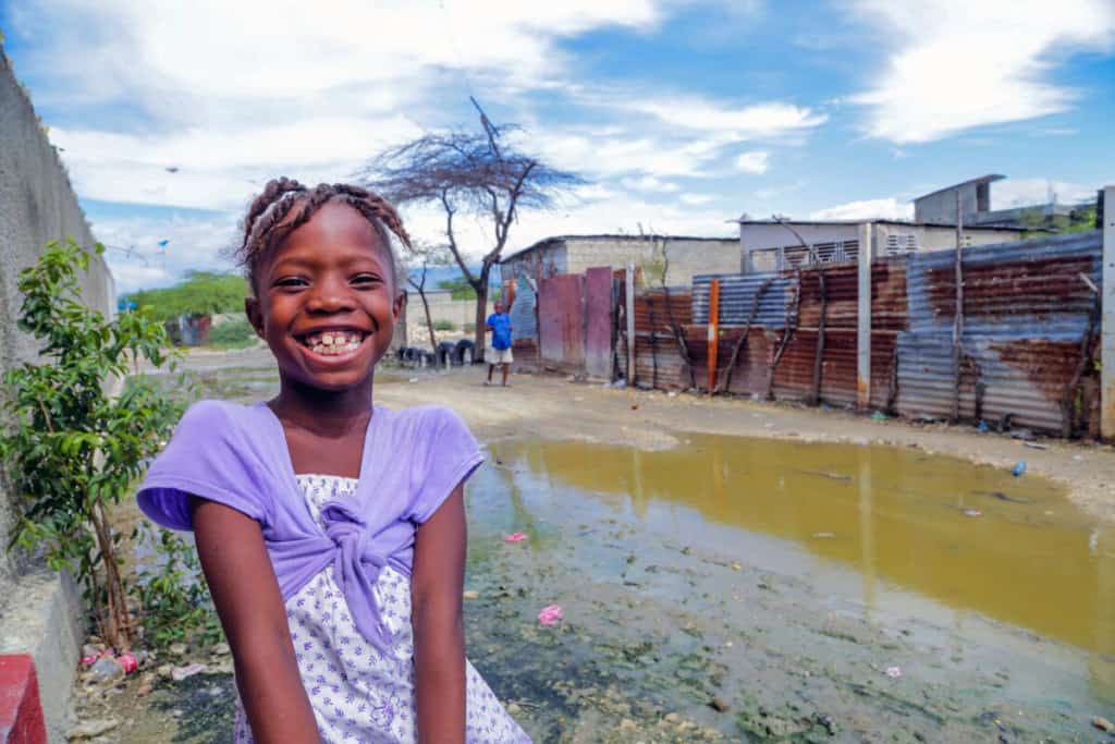 A smiling girl wearing a purple outfit sits in an impoverished neighborhood