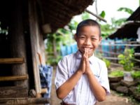 Suchat, a boy in Thailand, smiles and holds his hands in prayer. He is wearing a white collared shirt. A rural home and plants are seen behind him