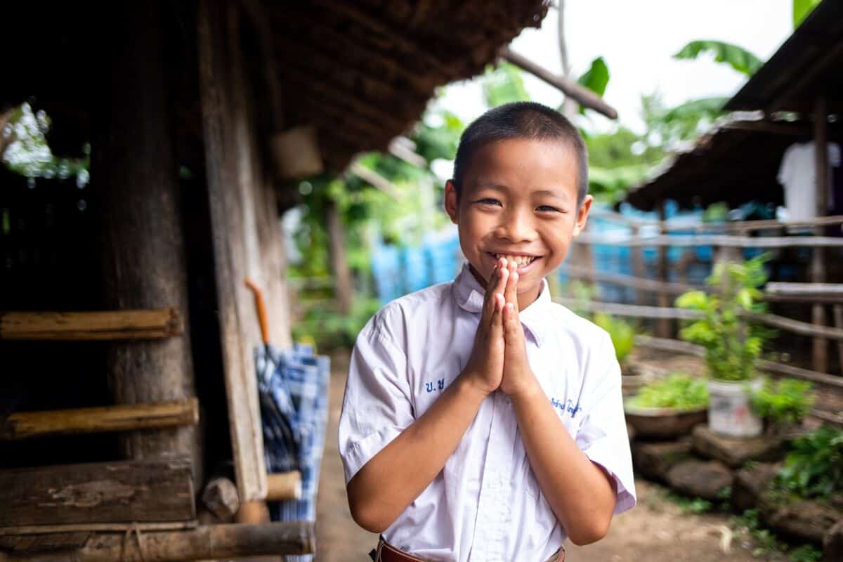 Suchat, a boy in Thailand, smiles and holds his hands in prayer. He is wearing a white collared shirt. A rural home and plants are seen behind him